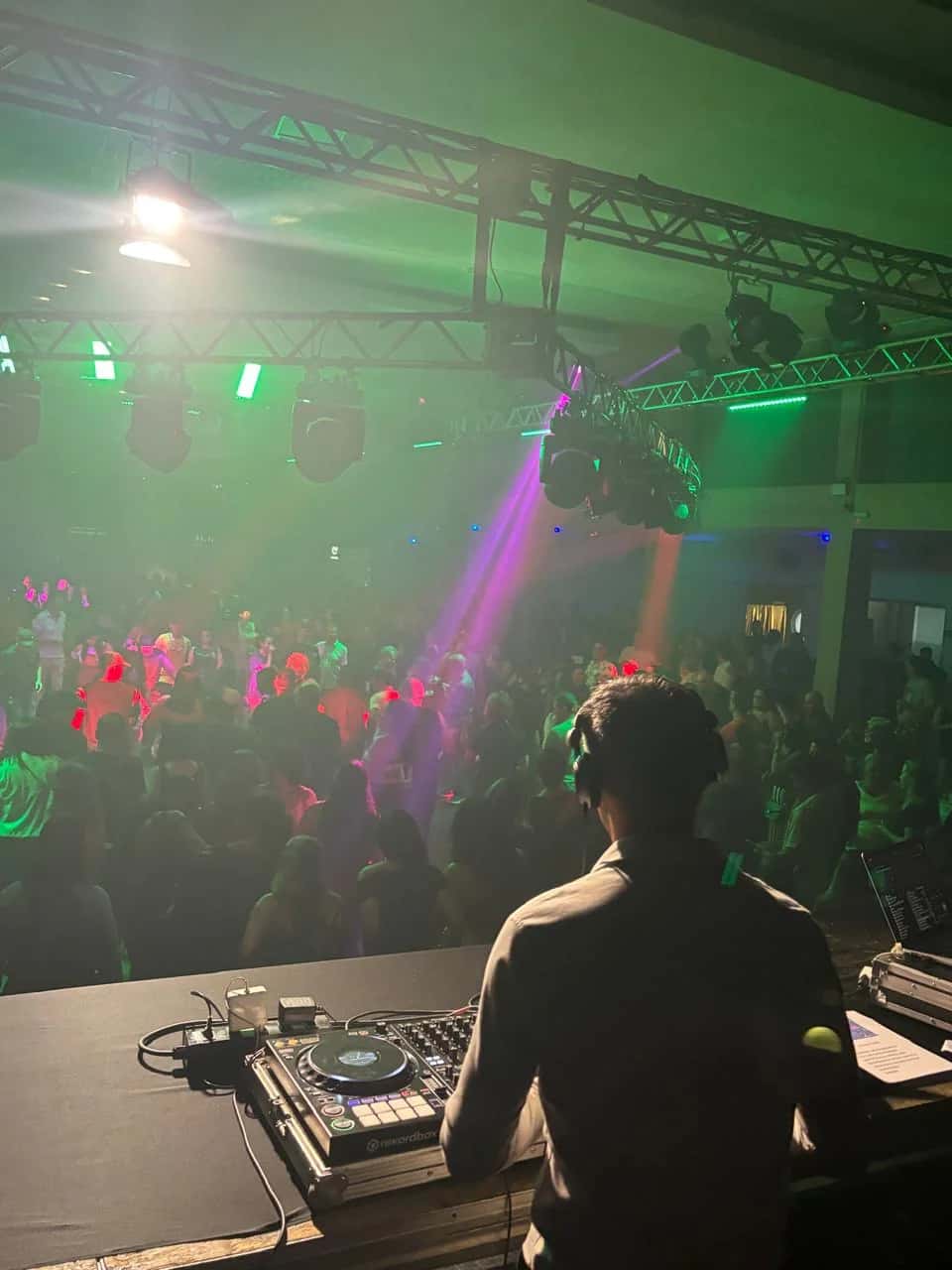 Vista do palco do DJ BetoBrizz para o público dançando sob luzes verdes e moving heads